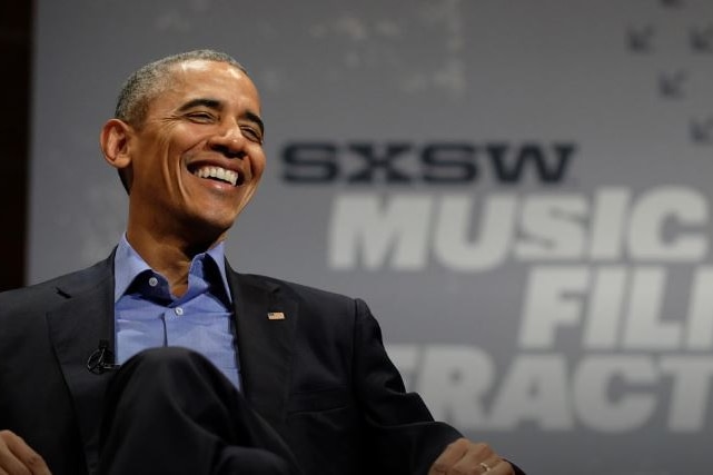 Barack Obama sitting down and smiling in front of a SXSW sign