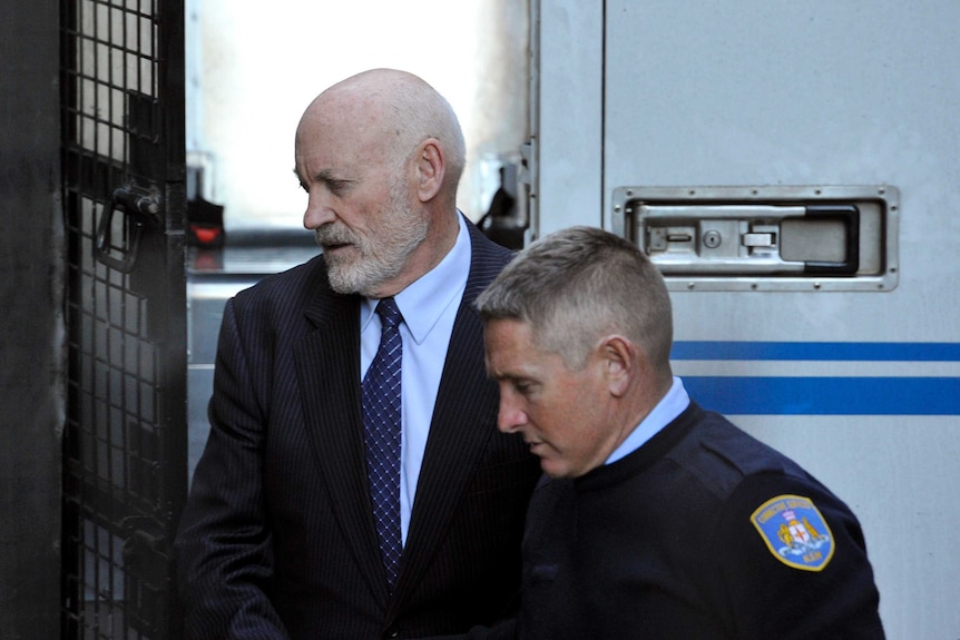 An older man is led away from a prison van by a police officer.