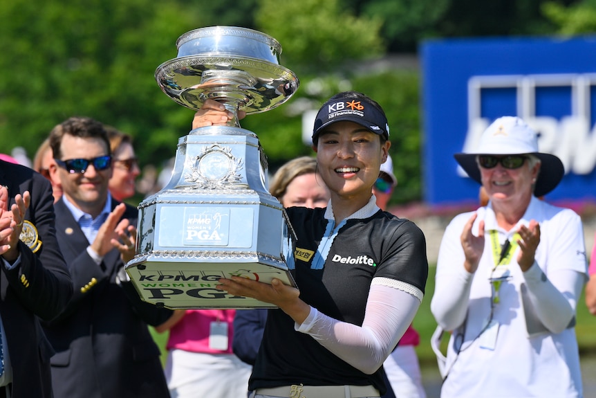 A female golfer smiles and accepts the crowd's applause as she holds up a very big trophy.