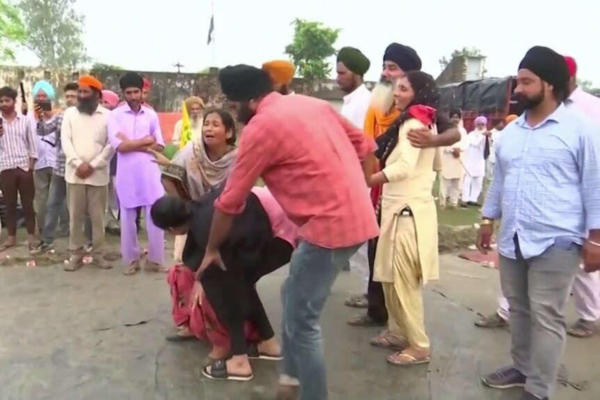 A South Asian family reacts emotionally on a footpath in a chaotic rural scene with people behind them.