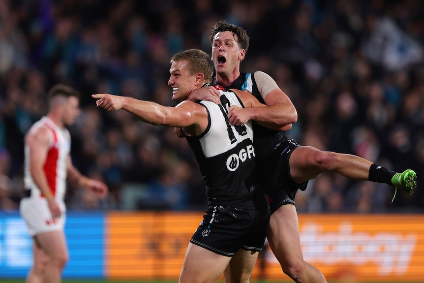 Two grinning Port Adelaide players hold on to each other in celebration as one points to a teammate.