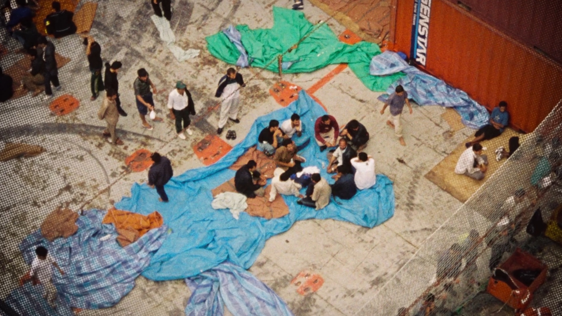 An aerial shot of people sitting on tarpaulins in the concrete deck of a ship.