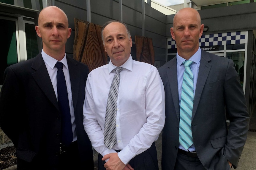 Three detectives stand side-by-side in wearing business attire in front of a station