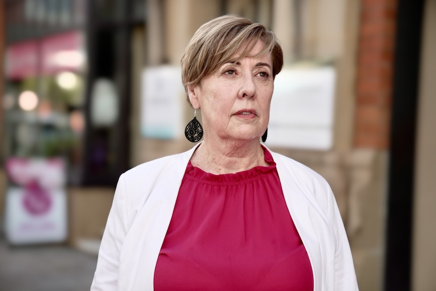 A woman looking off camera to her left. She has short, fair hair and is wearing a red top, white jacket & black earrings