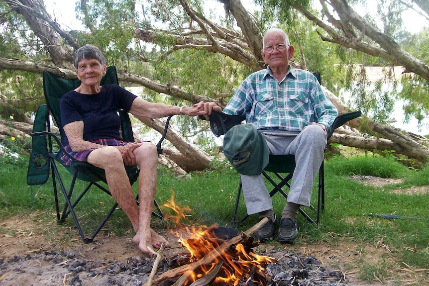 An elderly woman and man hold hands while sitting on camping chairs in the bush.