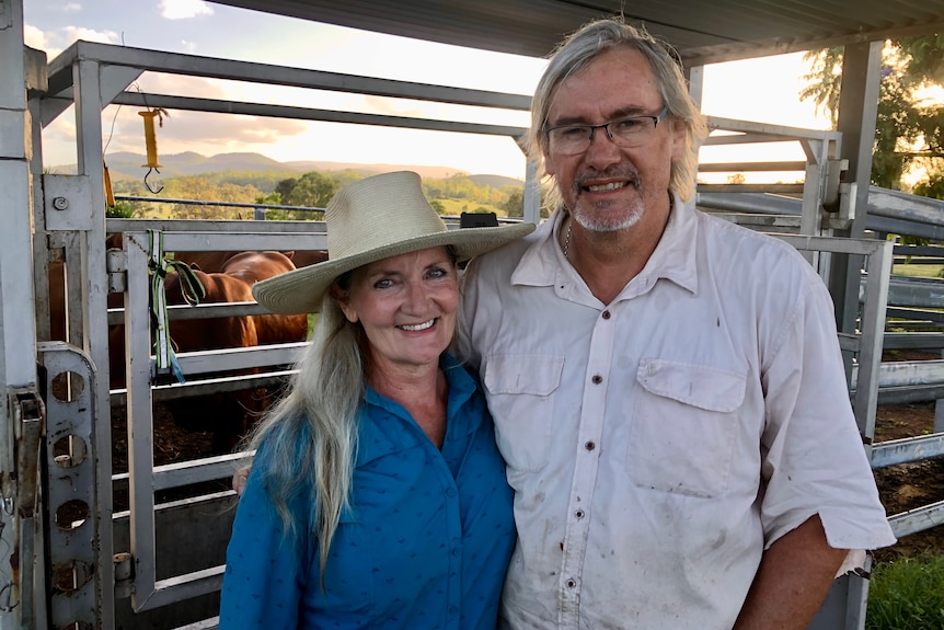 Female and male stand next to eachother in front of a yard where horses are being housed. 