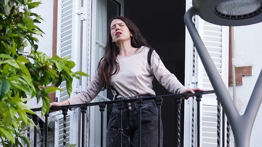 A woman wearing tan shirt and black pants, with long black hair, at a balcony with closed eyes and open mouth as if singing.