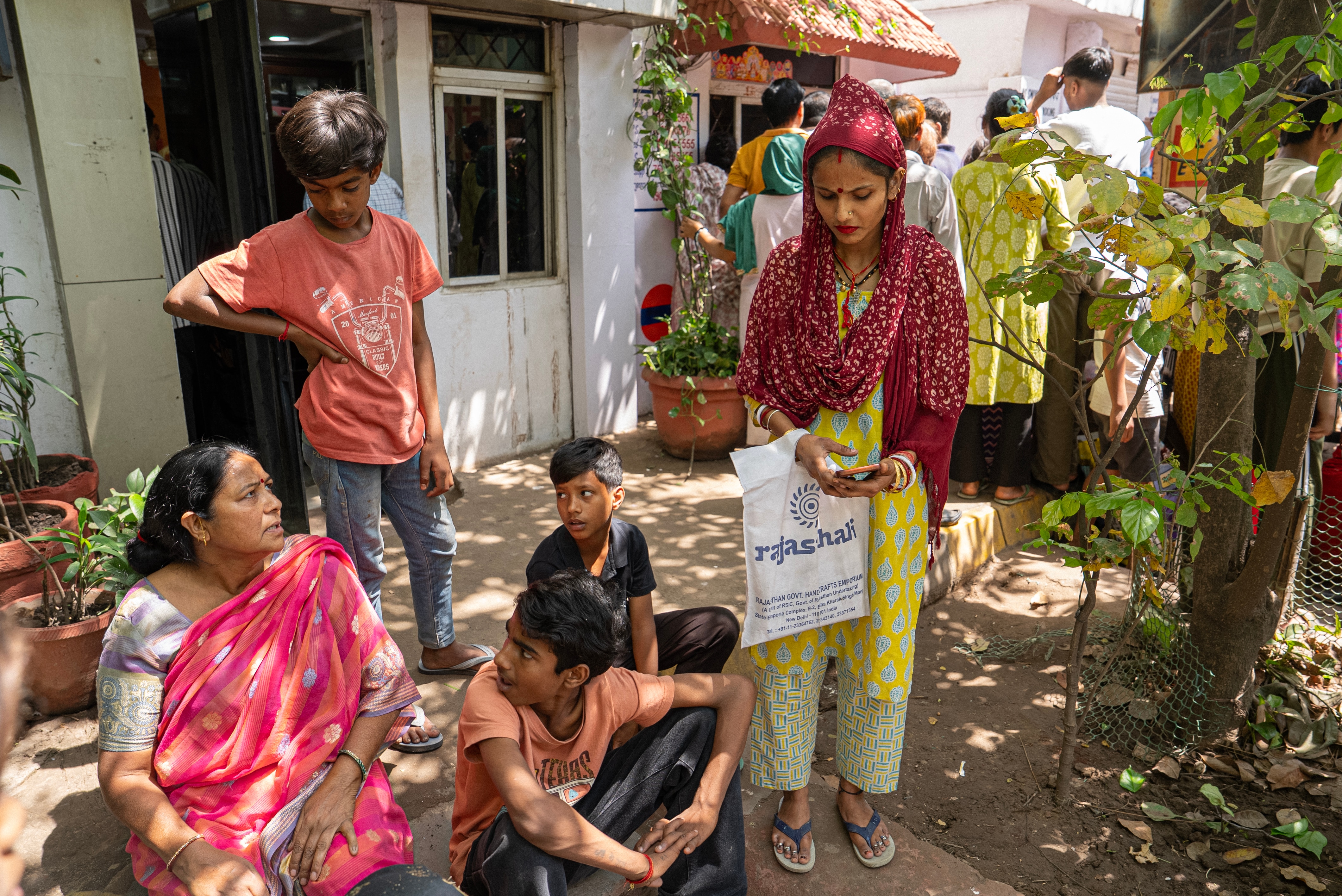 A woman sits on the ground looking at children, she wears a pink saree. Another woman, her covered in red stole, stands near.