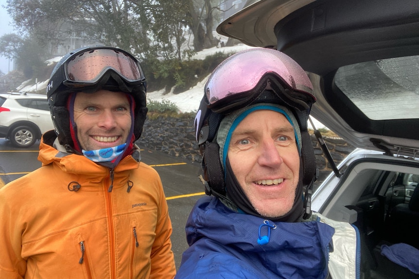 Two men stand at the boot of a car, wearing snow gear and smiling for a selfie.