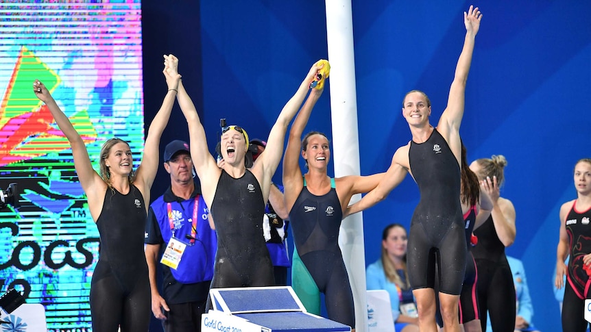 Four female swimmers in black swim suits raise their hands in the air and cheer after a win