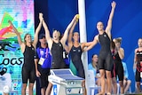 Four female swimmers in black swim suits raise their hands in the air and cheer after a win