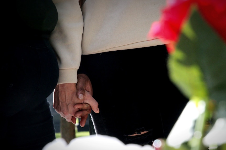 Two women hold hands with a blurry flower in the foreground.