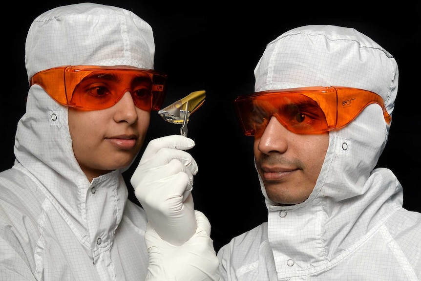 Professor Madhu Bhaskaran and Professor Sharath Sriram dressed in lab coats with orange safety goggles, looking at a metal