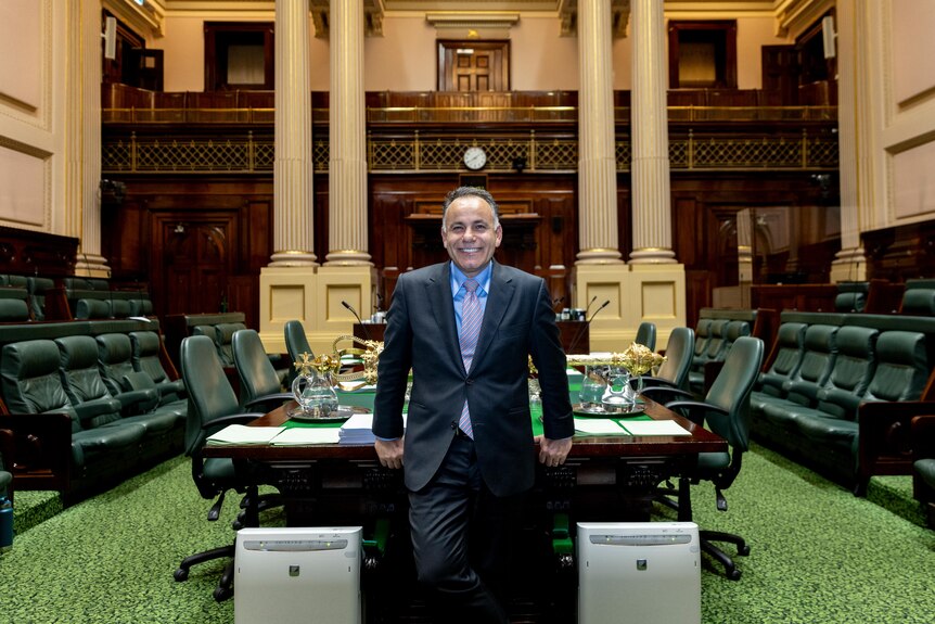 John Pesutto smiles, dressed in a suit and tie, standing in a green-carpeted chamber of parliament.