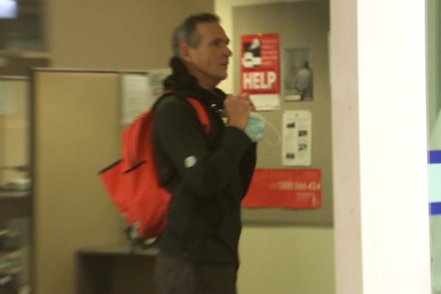 A man wearing dark clothes and a red backpack stands indoors, behind a glass sliding door