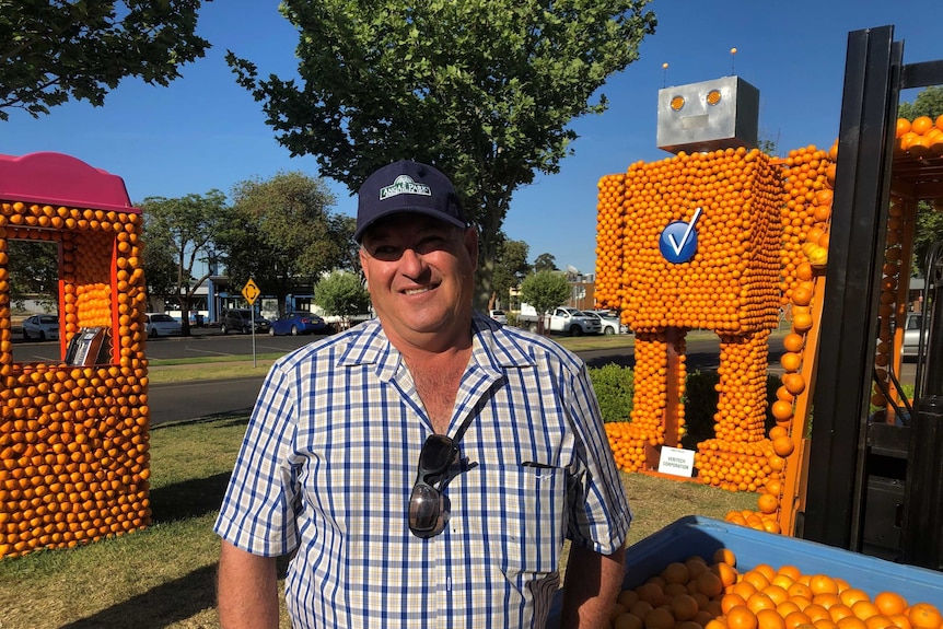 A man in a cap and chequered shirt smiles in front of carnival-like structures, including a giant robot, made of oranges.