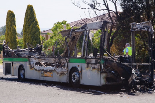 A bus sits on the side of the road destroyed by fire, with the roof and sides completely gone.