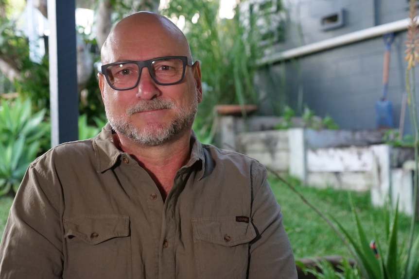 A man with black-framed glasses and a khaki, collared shirt looks at the camera.