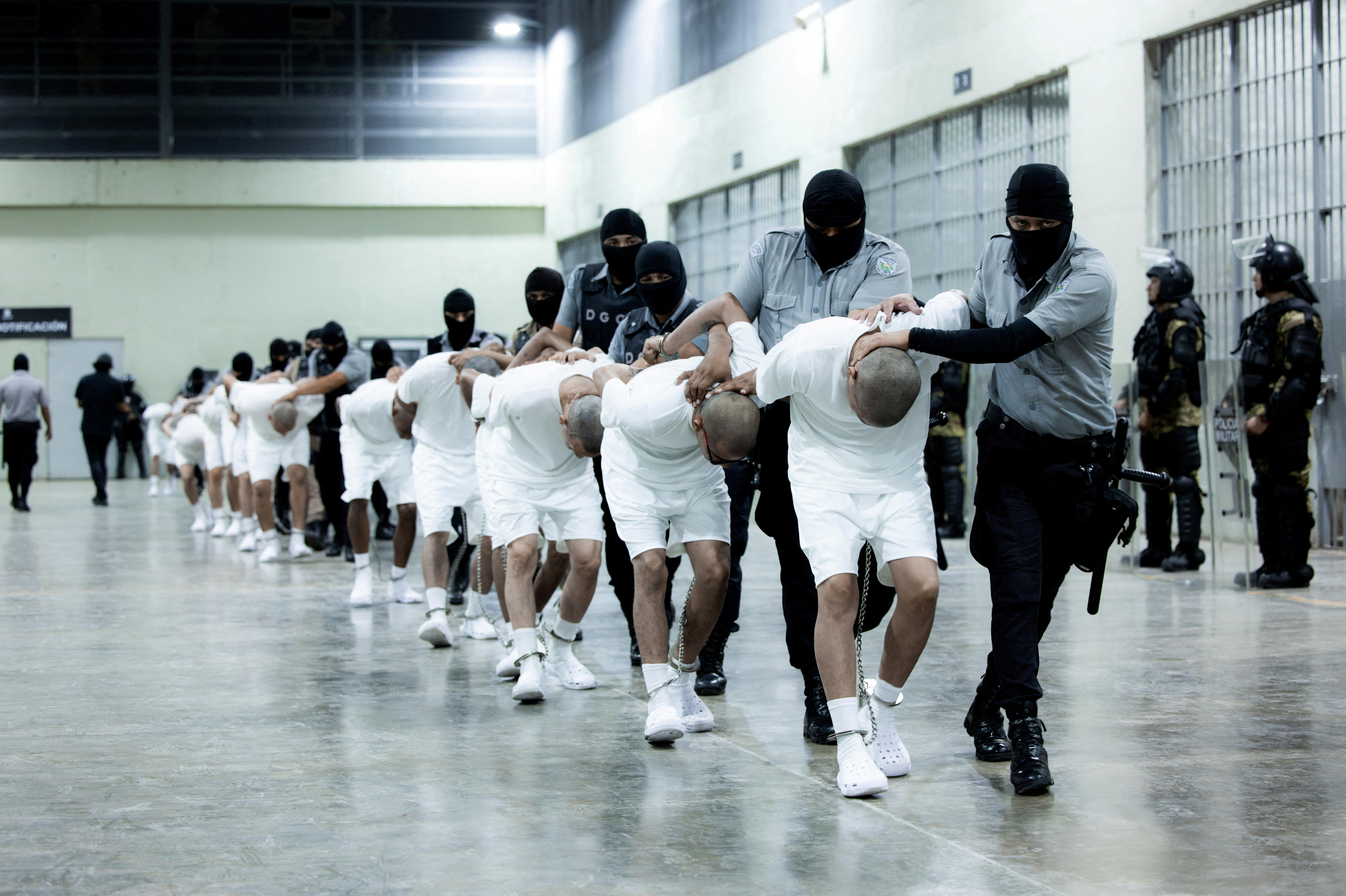Men in white suits are bent over and marched through a brightly lit prison.