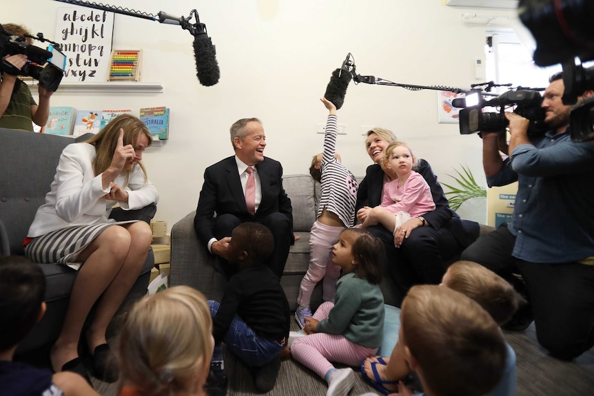 The politicians smile as the children sitting at their feet reach up to touch a boom microphone hovering above