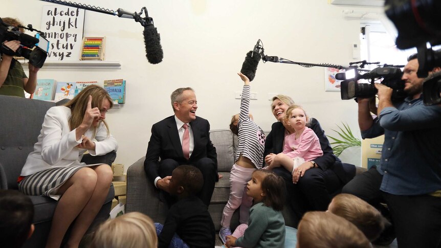 The politicians smile as the children sitting at their feet reach up to touch a boom microphone hovering above