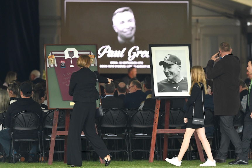 A screen showing Paul Green's face as funeral attendees arrive.