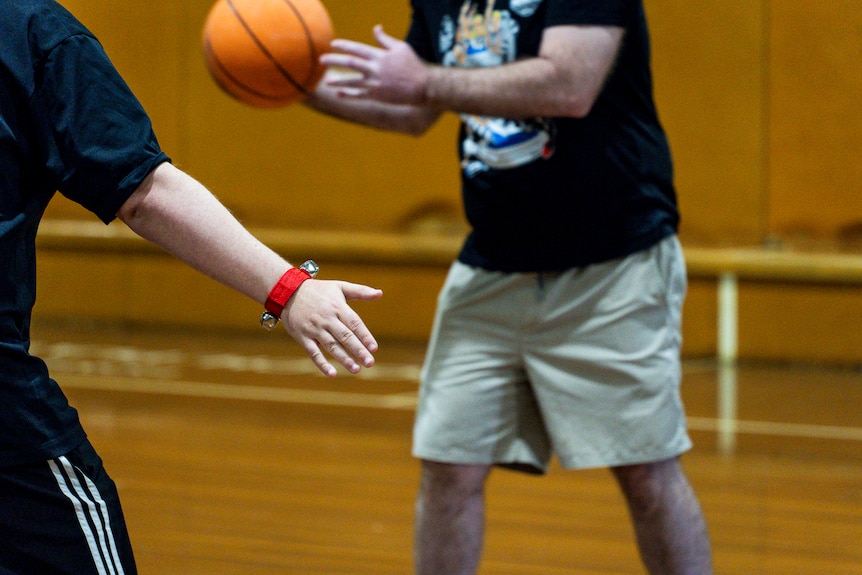 An outstretched hand with a wrist bell in the foreground, and a person holding a basketball in the background.