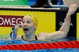 A woman in a pool wearing a yellow cap and holding blue goggles looks up in disbelief.