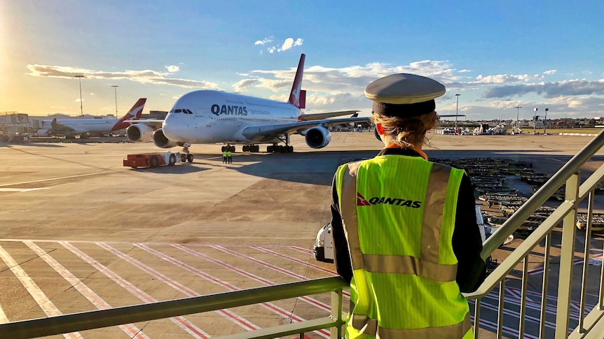 A pilot in a cap and high-vis vest looks from a platform at a Qantas A380 at an airport in the sunrise.