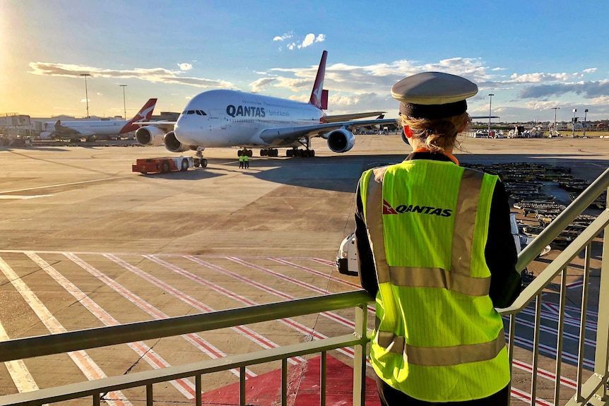 A pilot in a cap and high-vis vest looks from a platform at a Qantas A380 at an airport in the sunrise.