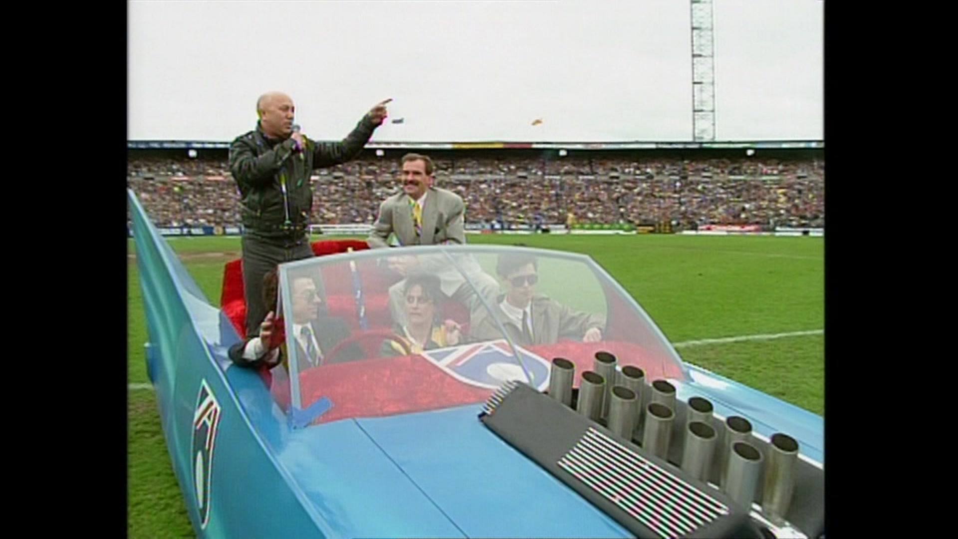 A bald man in a black leather jacket stands in the red seat of a light blue open air car pointing to a crowd in a stadium.