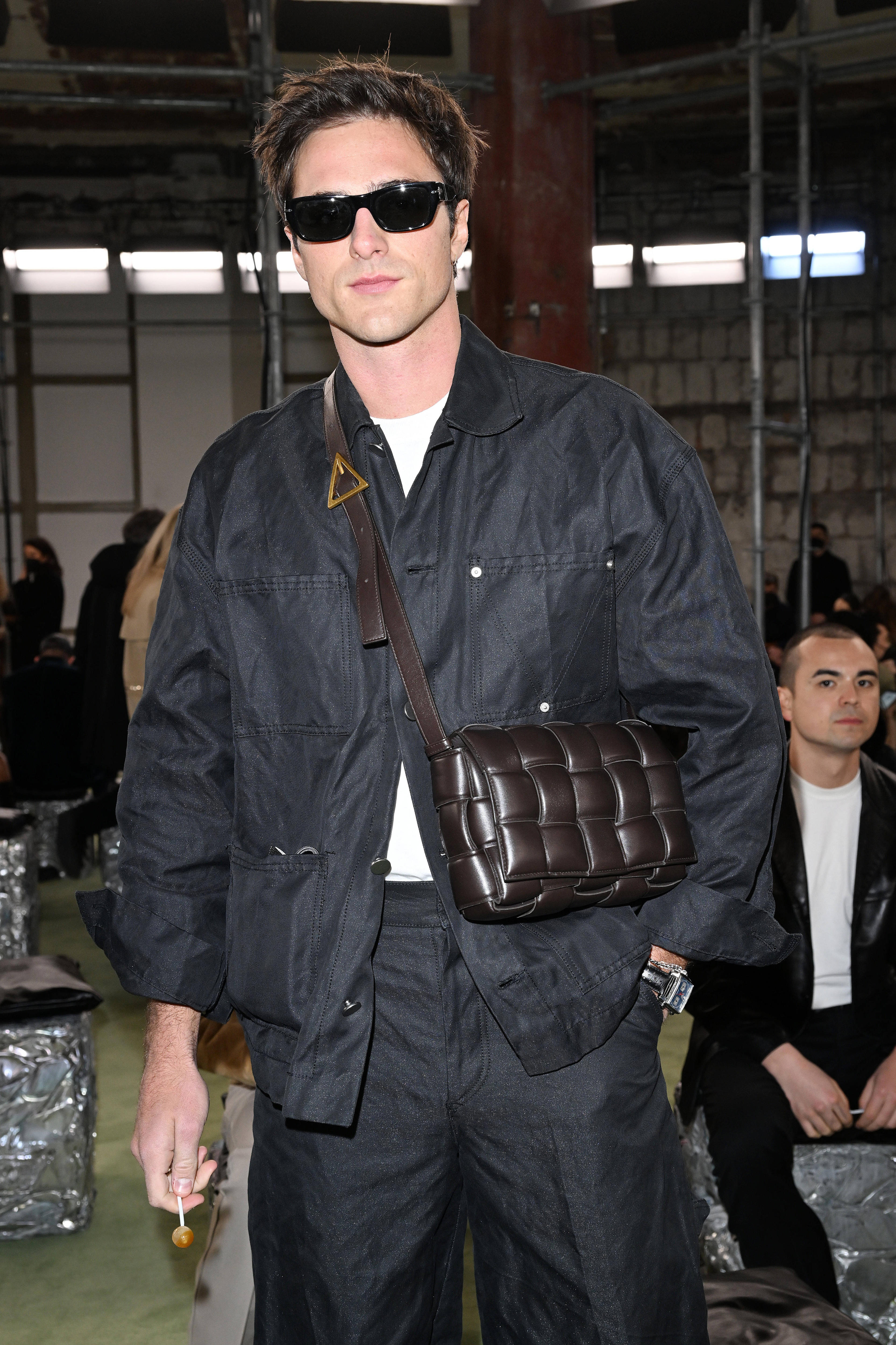 Jacob wears black sunglasses, a black jumpsuit and accessorises with a black leather crossbody handbag