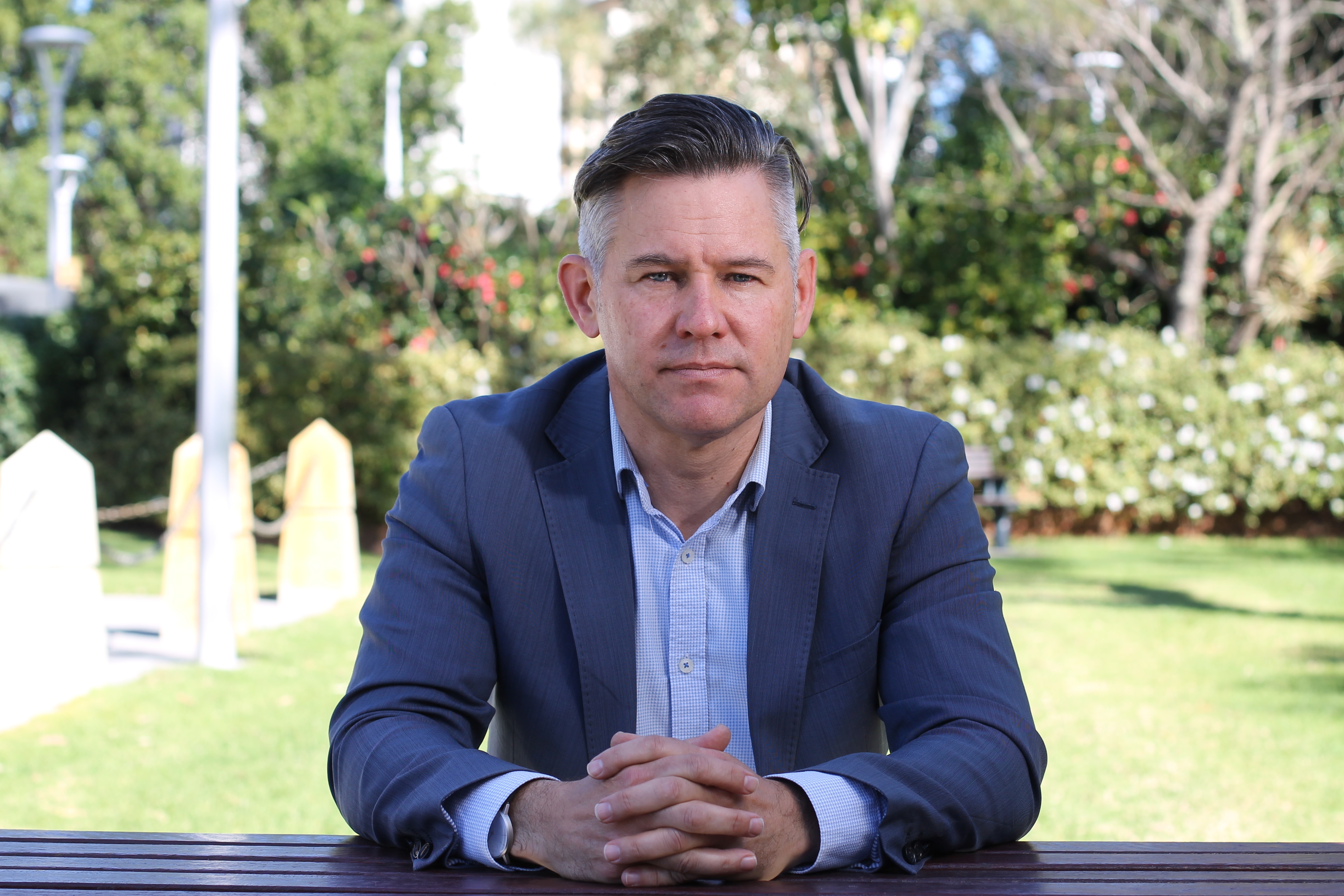 Close-up of Brad Pettitt, hands clasped, sitting on a park bench, looking at the camera.