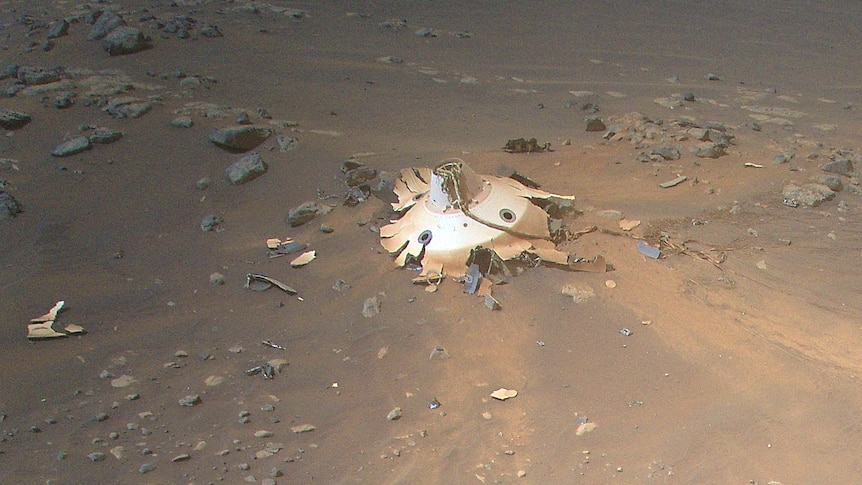 A colour image of the wreckage of a bronze metal pod laying on light brown dirt.
