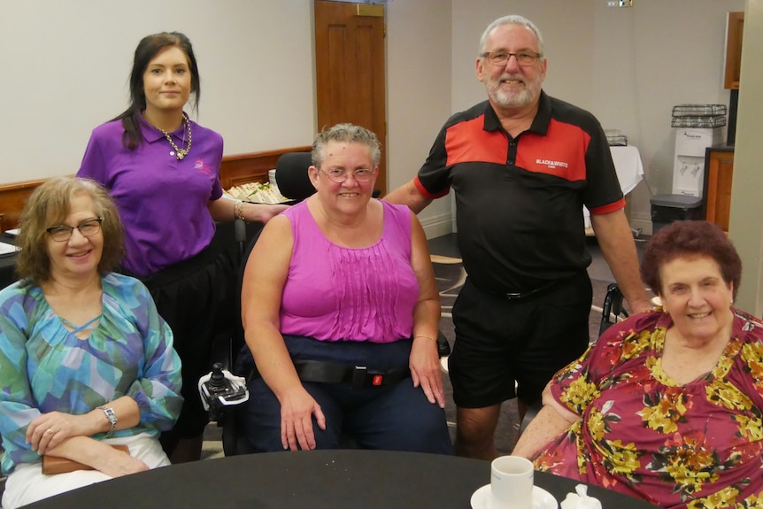 A man and four women smiling while they sit and stand around a table in a function room.