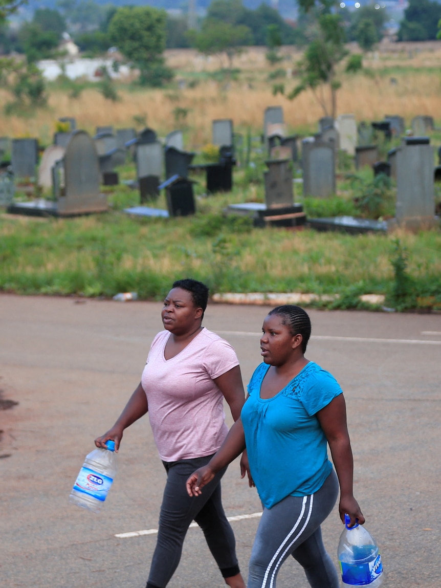 two women walk along a road inside a cemetery in Zimbabwe while holding drinking bottles