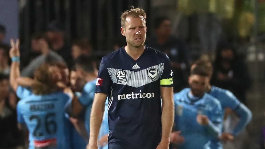 Melbourne Victory player Ola Toivonen walks towards the camera as Sydney FC players celebrate in the distance behind him.
