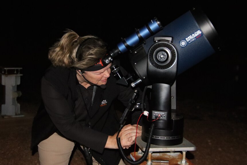 A woman wearing a head torch and dark jacket looking through a telescope on a patch of dirt at night.