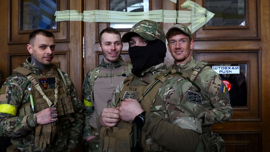 Four men in combat fatigues stand together, one with a British union flag patch on his chest.