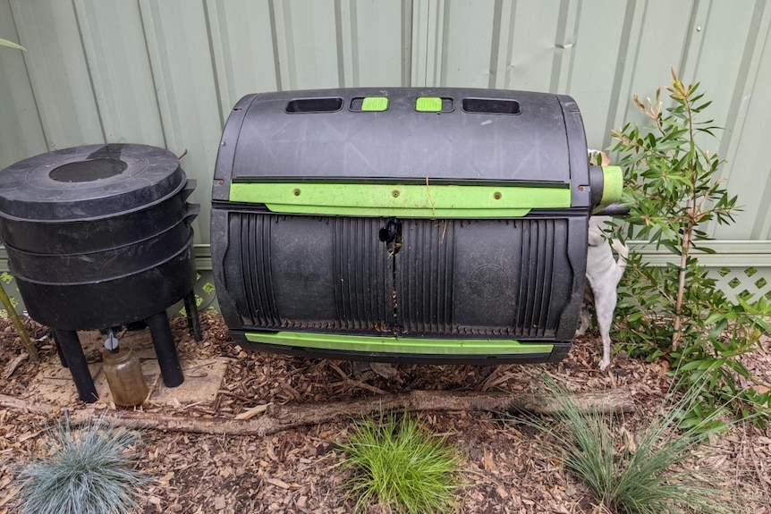 A black and green plastic compost tumbler stands in a backyard against a fence
