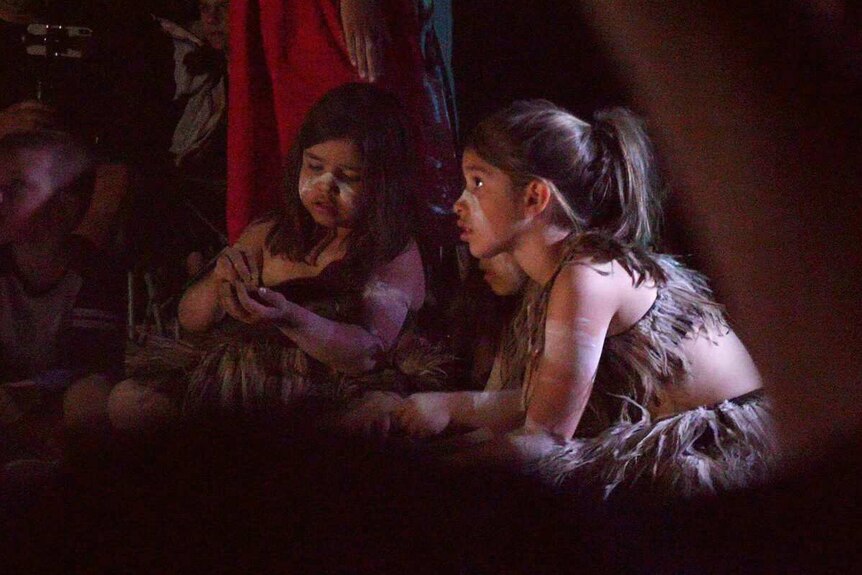 Two Aboriginal girls in emu feathers and painted faces sitting on the ground.