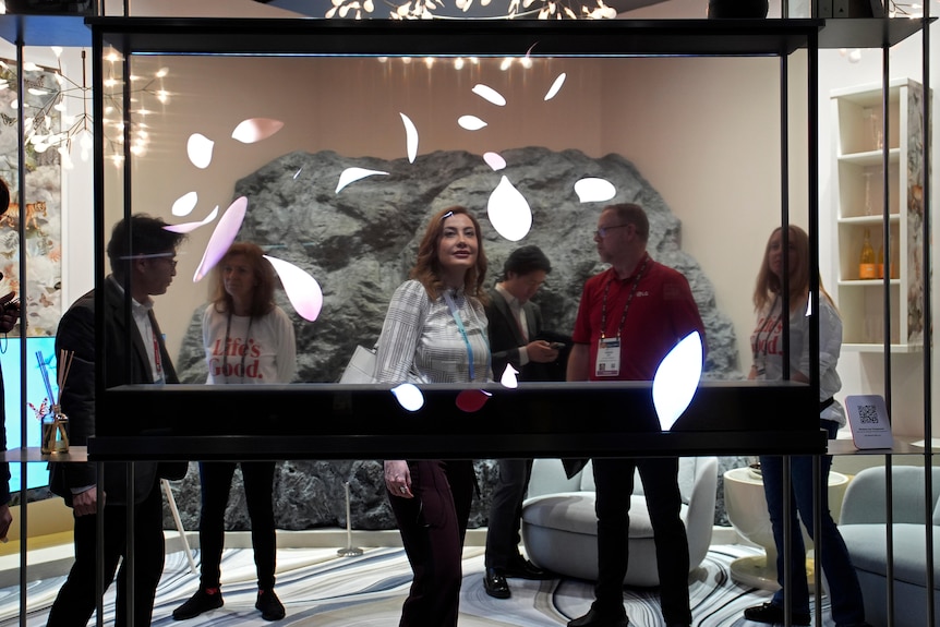 A medium shot of six people standing behind a transparent TV at a trade show, with some looking at the display