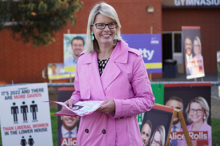 A blonde woman wearing a pink jacket holding how to vote cards