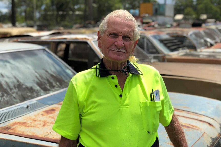 A man with white hair, wearing a hi-vis shirt, standing in front of a wrecking yard.