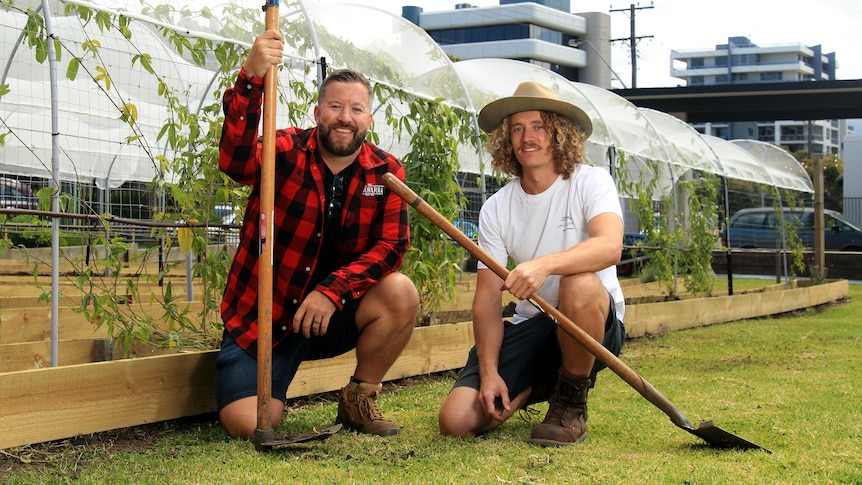 Ryan and Jared kneel down and hold shovels among their urban farm with passionfruit growing behind.