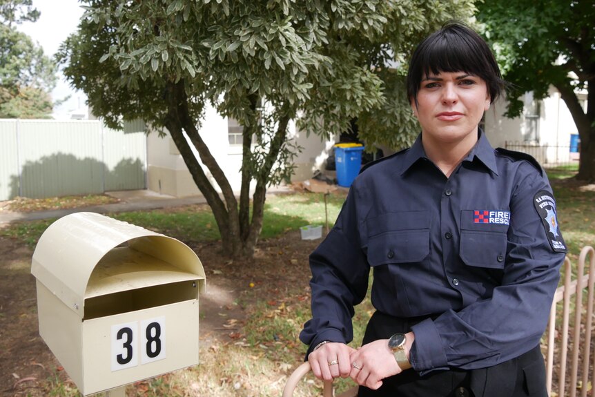 A woman with black hair and a fringe stands next to a letter box with a house behind her