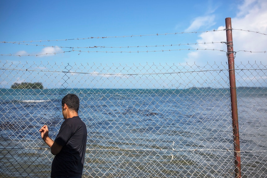A man with black hair and a black t-shirt leans against a tall wire fence, looking at the ocean