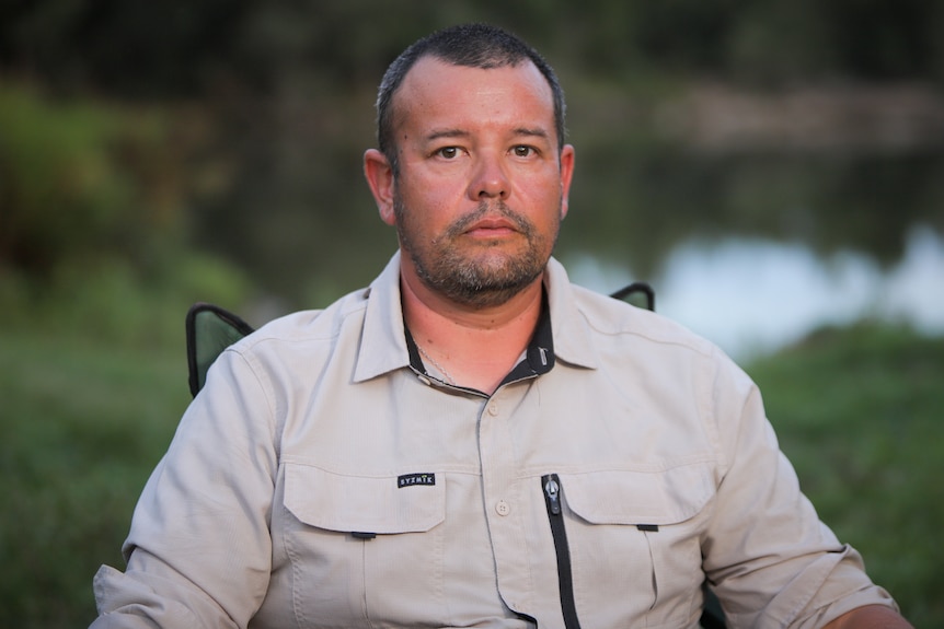 Man with shaved head wearing a khaki shirt sitting in a camping chair with a river behind him, looking serious.