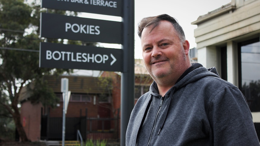 A middle aged man in a grey hoodie stands smiling outside a pokies venue