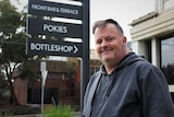 A middle aged man in a grey hoodie stands smiling outside a pokies venue